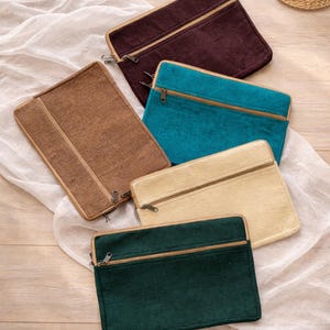 May include: A collection of rectangular zippered pouches in various colors: brown, teal, beige, dark green, and burgundy. Each pouch has a contrasting zipper and trim. The pouches are arranged on a white fabric.
