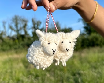 May include: Two small, fluffy white sheep ornaments with black eyes and a stitched smile, hanging from a red and white string. The sheep are made of a textured material, possibly wool, and are set against a blurred green field and blue sky.