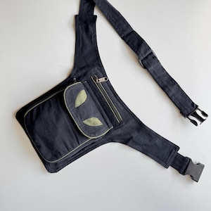 Puede incluir: Una riñonera negra de lona con una cremallera verde y un parche verde en la parte delantera. La riñonera tiene una correa larga ajustable con una hebilla.