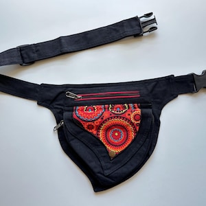 May include: Black fanny pack with a colourful, patterned front pocket. The pocket features a vibrant design with red, orange, and blue circular motifs. The pack has two zipped compartments and an adjustable black strap with a buckle.