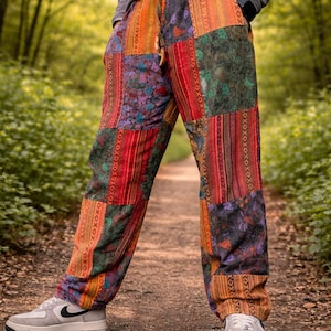 May include: Multi-coloured patchwork trousers with a loose fit and drawstring waist. The patchwork includes red, orange, green, and purple panels. The person is wearing grey trainers. The background is a blurred forest scene.