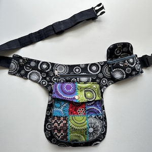 May include: A black waist bag featuring a white circular pattern, a central pocket with a colourful patchwork design, a smaller side pouch with a snap closure, and an adjustable black strap with a plastic buckle for secure fastening around the waist.