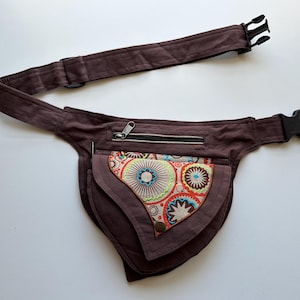 May include: A brown fanny pack with a decorative front pocket featuring a colourful circular pattern. The pack has a zipped top pocket and an adjustable strap with a black plastic buckle. The fanny pack is designed to be worn around the waist.