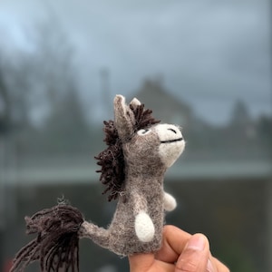 Puede incluir: Una figura de burro hecha a mano en fieltro, en tonos grises y marrones. El burro tiene una melena y cola marrón oscuro, detalles blancos en las pezuñas y una expresión sonriente. La figura está sostenida por una mano.