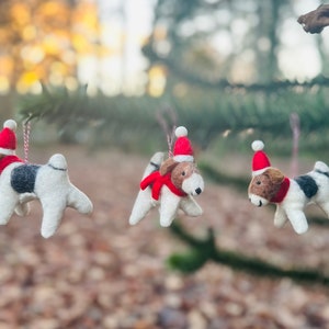 May include: Three felt Christmas ornaments shaped like dogs wearing Santa hats. The dogs are white with black spots and red and white scarves.