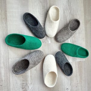 May include: Eight pairs of felt slippers in various colours, including grey, white, and green. The slippers are arranged in a circle on a light wood surface.