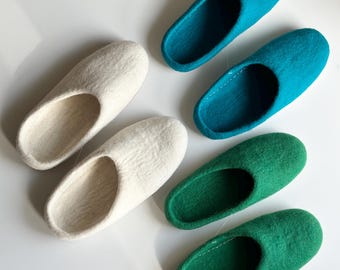 Puede incluir: Un conjunto de cuatro zapatillas de fieltro sin cordones dispuestas en pares. Dos zapatillas son de color blanco roto, mientras que las otras dos son de tonos verde azulado y verde. Cada zapatilla presenta una punta redondeada y abierta.