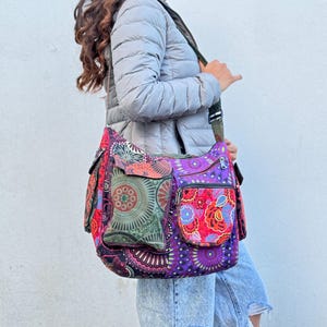 Op de afbeelding: Een kleurrijke schoudertas met meerdere vakken. De tas heeft een levendige mix van patronen in paars, rood, groen en oranje. Het heeft een lange riem om over de schouder te dragen. De tas is gemaakt van een duurzame stof.