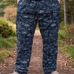 Könnte beinhalten: Dunkelblaue Hose mit einem sich wiederholenden Muster aus grauen Pilzen. Die Hose hat eine lockere Passform und wird mit beigen Schuhen getragen. Das Thema ist Natur und Botanik.