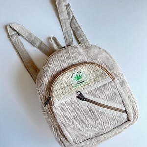 May include: A beige backpack featuring a herringbone pattern, adjustable shoulder straps, and a front zippered pocket. A rectangular label on the front pocket displays a green leaf icon and the text: HIMALAYAN HEMP THC FREE MADE IN NEPAL.