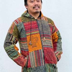 Könnte beinhalten: Eine Kapuzenjacke im Patchwork-Design mit Reißverschluss. Die Jacke hat eine Vielzahl von Farben, darunter Rot, Orange, Grün und Braun. Auf der Vorderseite befindet sich eine Pilz-Grafik. Die Jacke hat zwei Vordertaschen.