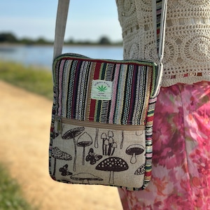 Puede incluir: Bolso bandolera rectangular de cáñamo. La parte superior es a rayas rojas, verdes y negras, mientras que la inferior presenta ilustraciones de setas y mariposas sobre un fondo beige. Tiene un bolsillo con cremallera y una correa ajustable.