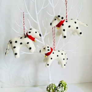 May include: Three white felt Dalmatian dog ornaments with black spots and red collars hanging from a white branch. Each ornament has a gold bell.