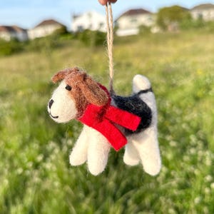 May include: A handmade felt dog ornament with a red scarf. The dog has brown and white fur with black accents. It is suspended by a natural twine string, perfect for hanging. The background is a blurred green field.