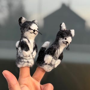 May include: Two handmade finger puppets shaped like black and white cats. The puppets are made of felt and have detailed features, including whiskers and small ears. One cat is upright, and the other is lying down. The puppets are on a person's fingers.