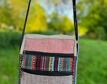 May include: A rectangular crossbody bag featuring a multi-colored woven fabric with a herringbone pattern. The bag has three horizontal zippered compartments, a black adjustable strap, and a small green label with a leaf icon and text.