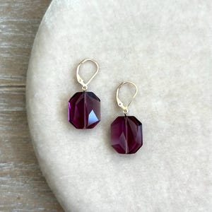 May include: A pair of faceted, deep purple crystal earrings with silver-colored lever-back closures. The earrings have an octagonal shape and are displayed on a light-colored, textured surface. The earrings are a jewelry accessory.