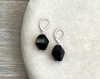 May include: A pair of black faceted gemstone earrings with silver-colored leverback closures. The earrings have a geometric, angular design, and the gemstones are a deep, glossy black. The earrings are displayed on a light-colored, textured surface.