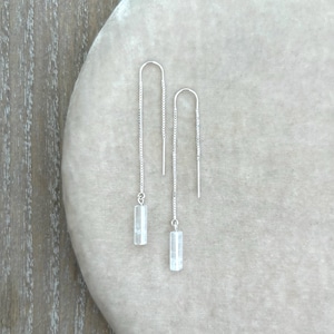May include: A pair of silver threader earrings with rectangular, clear crystal pendants. The earrings are displayed on a light gray surface, showcasing their delicate design and minimalist style. The earrings are a modern accessory.