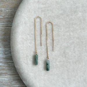 May include: A pair of gold-tone threader earrings featuring delicate box chains. Each earring terminates in a small, cylindrical, mottled green stone bead. The design consists of a long, thin metal chain that loops through the earlobe.