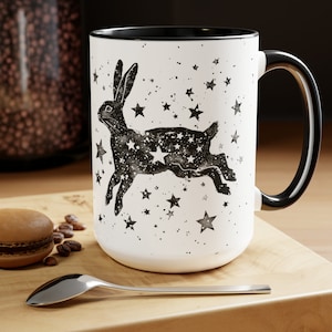 Puede incluir: Taza de cerámica blanca con asa y borde negros. La taza presenta una ilustración en blanco y negro de un conejo con estrellas dentro de su cuerpo. El conejo está rodeado de estrellas más pequeñas.