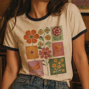 Può includere: T-shirt color crema con un design floreale retrò. La maglietta ha un bordo blu navy attorno al colletto e alle maniche. Il motivo floreale presenta fiori arancioni, rosa, gialli e verdi in una griglia.
