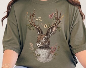 Könnte beinhalten: Olivgrünes T-Shirt mit einer detaillierten Illustration eines Hasen mit Hirschgeweih, umgeben von Blumen und Blättern. Das Kunstwerk ist auf dem T-Shirt zentriert und schafft ein einzigartiges und künstlerisches Design. Das Shirt hat kurze Ärmel und eine lockere Passform.
