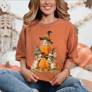 May include: Burnt orange t-shirt featuring a whimsical illustration of three white rabbits wearing witch hats, perched on pumpkins. The design has a fall or Halloween theme, with detailed artwork and a cozy aesthetic. The shirt is a comfortable, casual style.