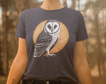 May include: A navy blue t-shirt with a white and black barn owl illustration. The owl is surrounded by a gold sunburst design.