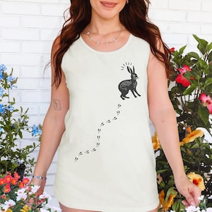 Puede incluir: Camiseta sin mangas color crema con una ilustración de conejo negro, líneas radiantes sobre su cabeza y un rastro de huellas. La camiseta sin mangas es de estilo casual, adecuada para climas cálidos.