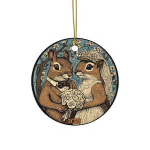 Op de afbeelding: Ronde keramische ornament met een eekhoornkoppel in trouwkleding. De mannelijke eekhoorn draagt een smoking en een vlinderdas, de vrouwelijke eekhoorn een sluier en houdt een boeket vast. De achtergrond is een blauwe lucht met bloeiende takken. Het jaar "2025" staat onderaan gedrukt.