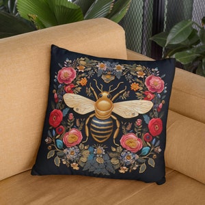 Puede incluir: Una almohada negra con una abeja dorada rodeada de flores coloridas. La abeja tiene rayas negras y amarillas y est&aacute; rodeada de una corona de flores en tonos rojos, rosas y azules.