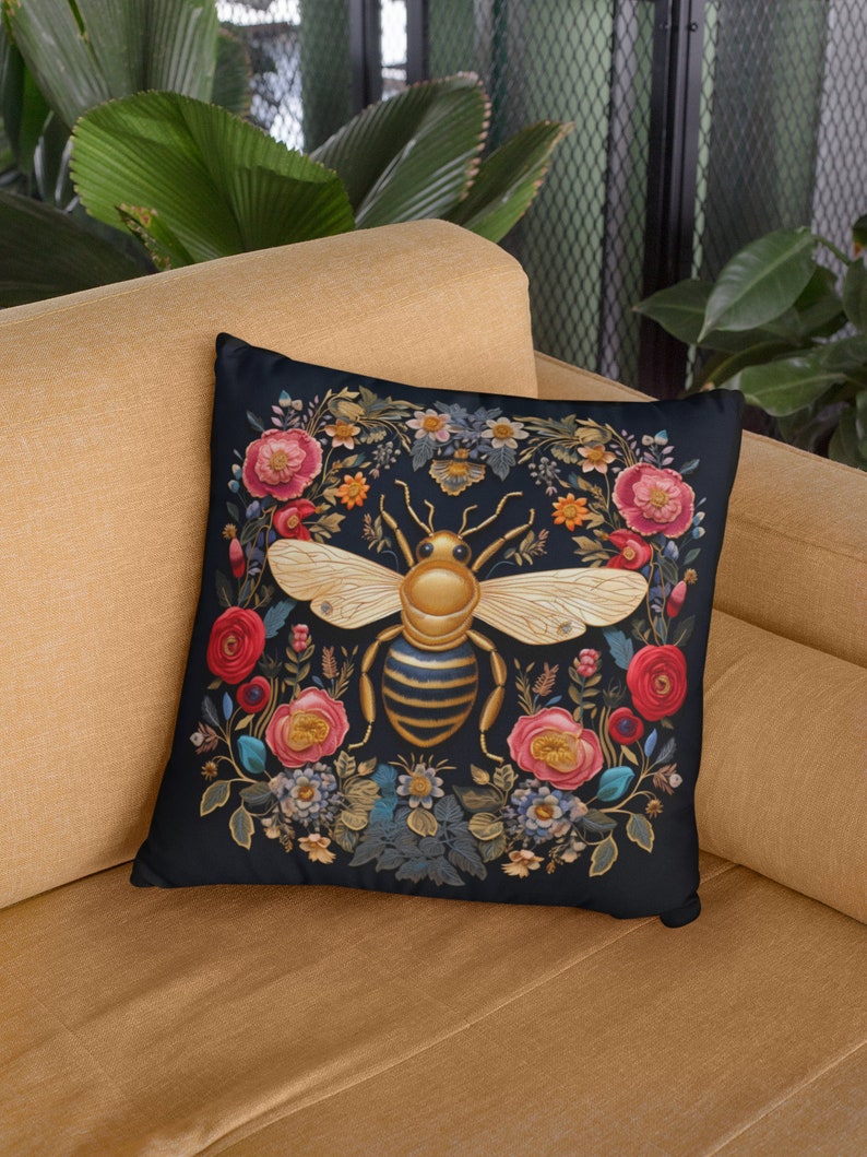 Puede incluir: Una almohada negra con una abeja dorada rodeada de flores coloridas. La abeja tiene rayas negras y amarillas y est&aacute; rodeada de una corona de flores en tonos rojos, rosas y azules.