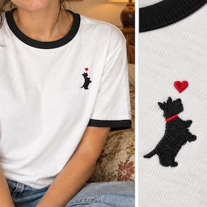May include: A white ringer t-shirt with black trim on the crew neckline and sleeve cuffs. The chest features a small embroidered black Scottish Terrier dog wearing a red collar, positioned below a single red heart stitched in thread.