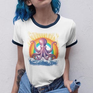 May include: White ringer t-shirt with a navy blue trim featuring a purple octopus design set against a rainbow and ocean waves. The octopus has multiple tentacles and is centered on the shirt.