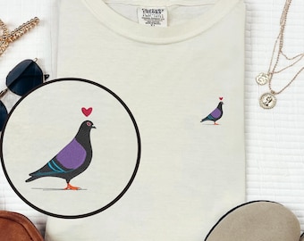 May include: Cream-colored t-shirt featuring an embroidered pigeon design with a heart above its head. The pigeon has a purple body, black wings, and red feet. The shirt has a classic crew neck and a relaxed fit, perfect for casual wear.
