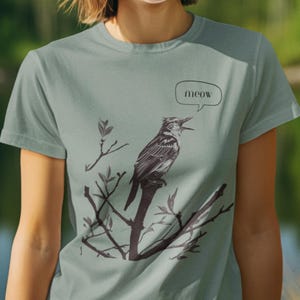 Puede incluir: Camiseta de color verde azulado claro con una ilustración en blanco y negro de un pájaro posado en una rama. Una burbuja de diálogo sobre el pájaro dice "meow". La camiseta es de cuello redondo y manga corta.