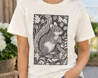 May include: A cream-colored short-sleeve t-shirt featuring a black graphic print of a squirrel surrounded by detailed botanical elements, including leaves and acorns, arranged in a rectangular frame on the front of the garment.