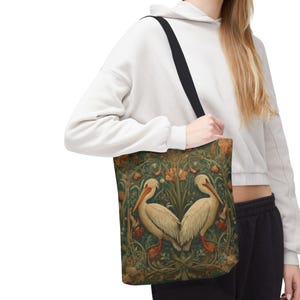 May include: A tote bag with a black strap featuring a vintage-style design. The bag displays two white pelicans facing each other, set against a backdrop of green foliage and orange flowers. The overall aesthetic is reminiscent of classic botanical illustrations.