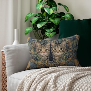 Könnte beinhalten: Rechteckiges Zierkissen mit einem symmetrischen Design von zwei Tabby-Katzen, umgeben von Blättern. Das Kissen hat einen dunkelblauen Hintergrund mit grünen, goldenen und braunen Akzenten. Das Kissen liegt auf einem hellgrauen Sofa mit einer cremefarbenen Decke.
