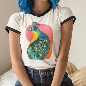 May include: White ringer t-shirt with a colorful peacock graphic. The peacock features teal, yellow, and orange hues against a pink backdrop. The shirt has a black trim around the collar and sleeves.