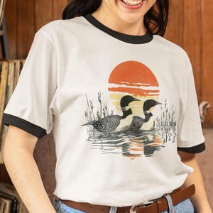 May include: White t-shirt with a black ringer neckline featuring a graphic of two loon birds swimming in a lake with a sunset in the background.