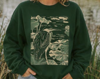 May include: A dark forest green crewneck sweatshirt featuring a light cream-colored graphic print of a heron standing among water lily pads. The long-sleeved pullover has a ribbed neckline, cuffs, and hem, showcasing a detailed nature illustration.