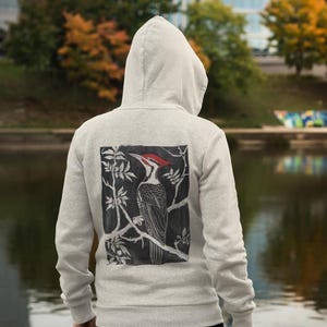May include: Light gray hooded sweatshirt with a graphic of a woodpecker perched on a branch. The woodpecker has a red head and is set against a black background. The sweatshirt is made of a soft material.