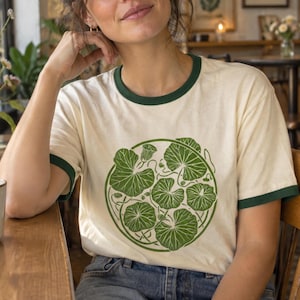 May include: A cream-colored ringer t-shirt with dark green trim on the collar and sleeve cuffs. The front features a circular graphic design composed of multiple green nasturtium leaves and a single small flower bud arranged in a cluster.