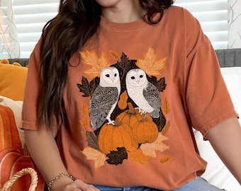 Peut inclure: Un t-shirt à col rond orange brûlé avec une impression graphique représentant deux effraies des clochers blanches perchées sur des citrouilles orange, entourées de feuilles d'automne détaillées dans des tons orange, jaune et noir.