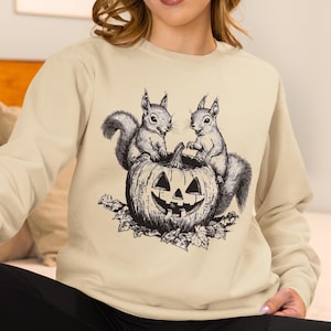 May include: A beige crewneck sweatshirt featuring a black ink-style graphic of two squirrels perched on a carved jack-o'-lantern pumpkin surrounded by fallen leaves. The design is centered on the chest of the long-sleeved pullover garment.