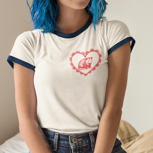 Puede incluir: Una camiseta blanca con cuello azul y un corazón rojo con un opossum dentro. El corazón tiene un diseño de encaje rojo.