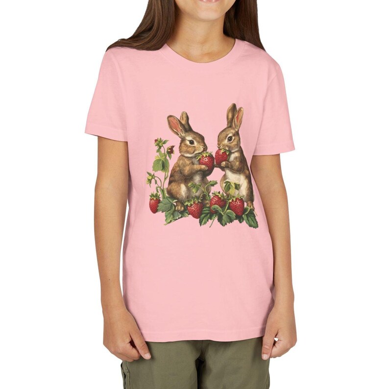May include: Pink t-shirt featuring a graphic of two brown bunnies eating strawberries. The bunnies are surrounded by strawberry plants with red berries and green leaves. The shirt is a light pink color.