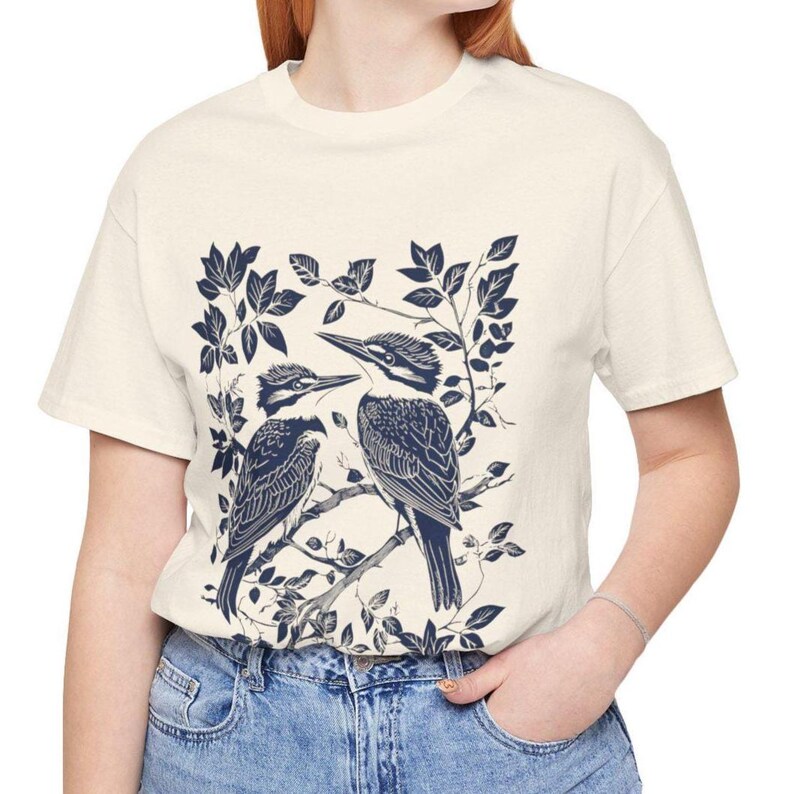 Op de afbeelding: Cr&egrave;mekleurig T-shirt met een donkerblauwe illustratie van twee vogels op een tak, omringd door bladeren. De vogels hebben gedetailleerde verenpatronen. Dit casual T-shirt is geschikt voor dagelijks gebruik.